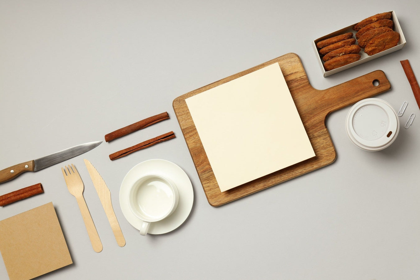 Flatlay mit Küchenutensilien, Schneidebrett, Tasse und Zimtstangen auf hellgrauem Hintergrund – stilvolle Darstellung für Haushalt & Küche.