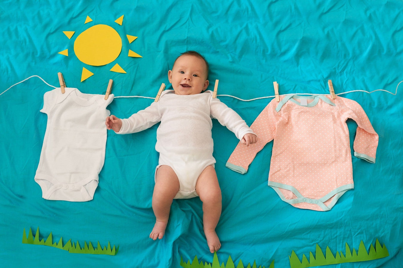 Baby liegt lachend auf blauem Hintergrund zwischen Kinderkleidung – süßes Flatlay mit Body & Accessoires für TECHTRAL Modekollektion
